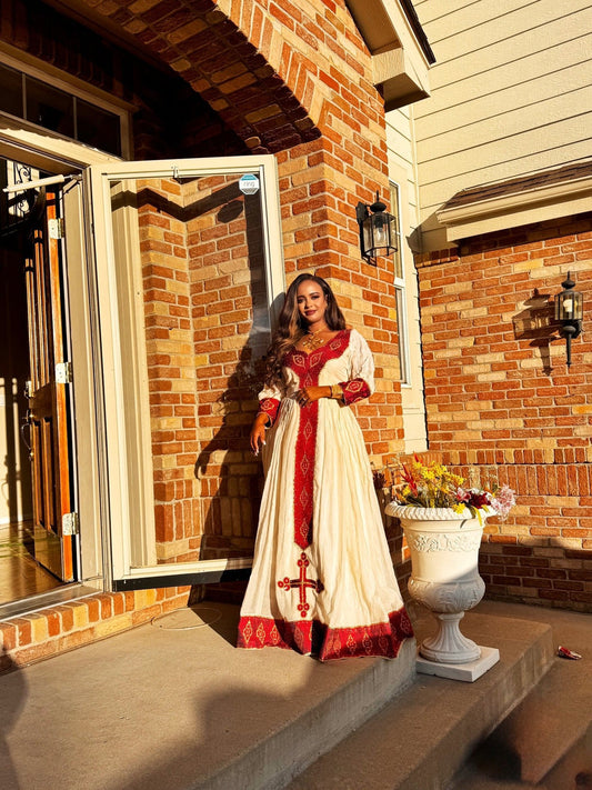 Woman in a white and red habesha kemis standing on a porch with a brick wall background