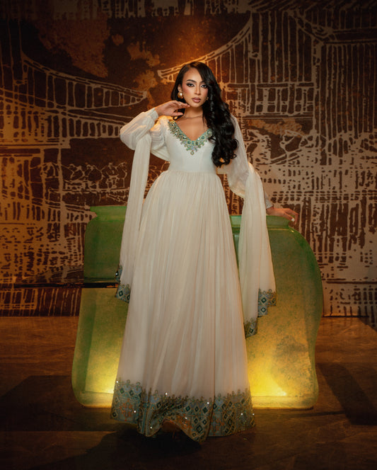 A woman models a long white habesha kemis with green and blue embellishments, standing in front of a stylized green backdrop against a dark patterned wall.