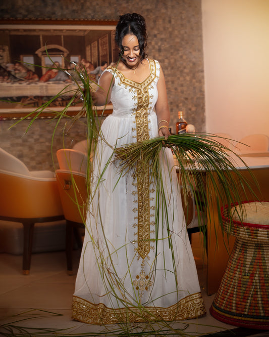 A woman wearing a white sleeveless Ethiopia Clothes with ornate gold embroidery and a gold border stands indoors while holding tall green grasses. The room has warm lighting, upholstered chairs, and a woven basket visible in the background.