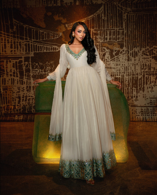 A woman models a long white or ivory traditional habesha kemis with turquoise and green embroidered detailing at the neckline, sleeves, and hem, standing in front of a dark illustrated backdrop.