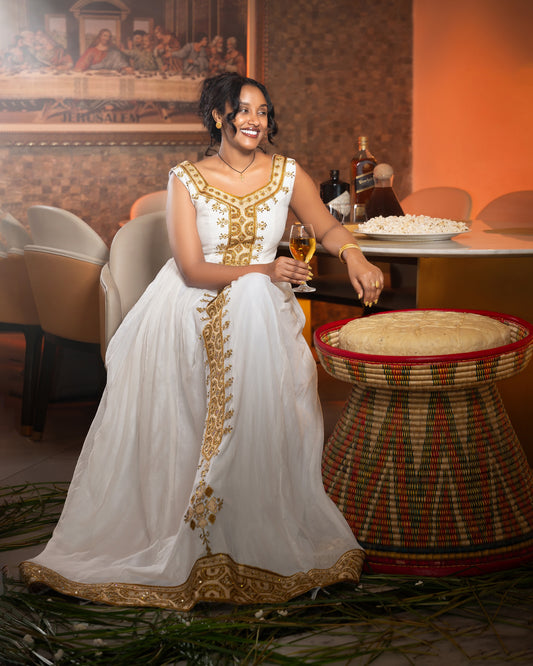 A woman in a white traditional Ethiopia Clothes with gold embroidery sits in a restaurant setting, holding a glass of champagne next to a woven table.