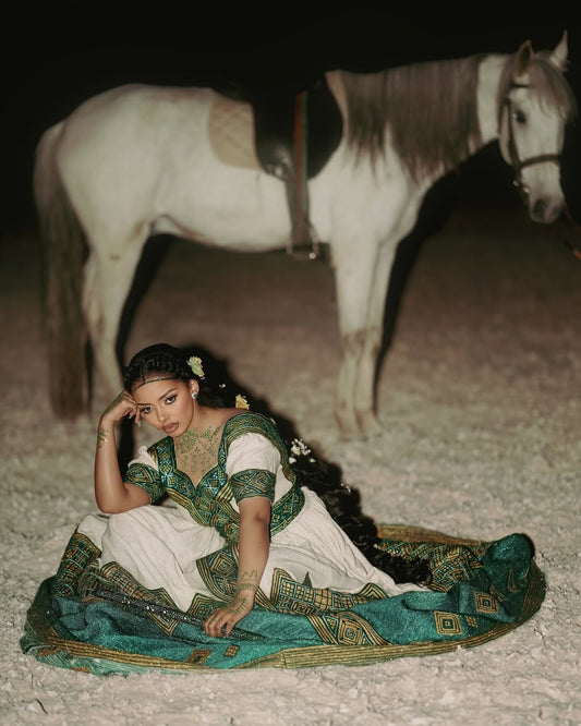 Woman in Ethiopian Habesha Wedding Dress attire sitting on the ground with a horse in the background