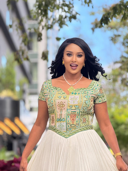 A woman wearing a traditional-style Habesha Dress with a green and beige patterned embroidered bodice and a white pleated skirt, standing outdoors with blurred trees and buildings in the background.