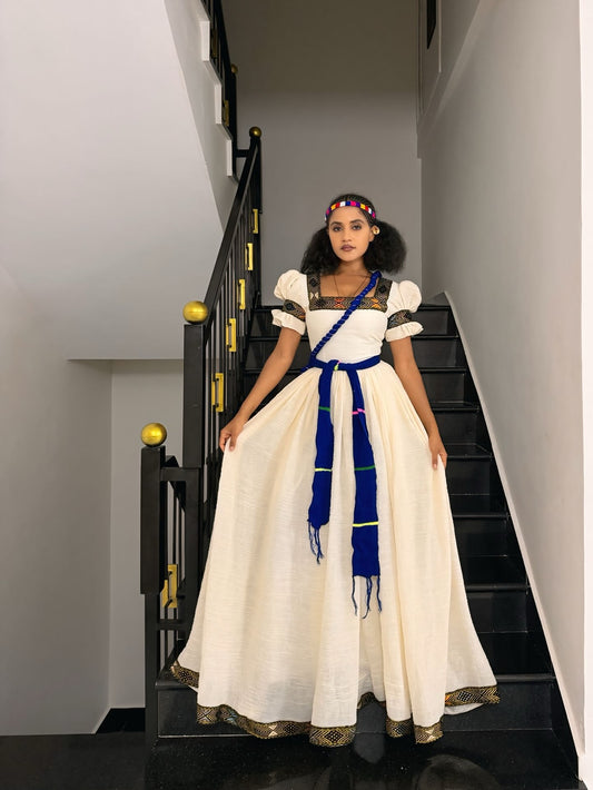 A woman stands on a black staircase wearing a traditional-style cream Ethiopian Dress with puffed sleeves, a blue sash, and gold trim, against a white wall.