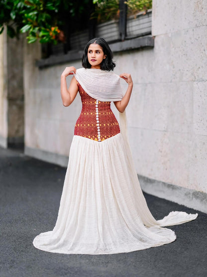 Woman wearing a traditional habesha kemis with a red blouse and white saree, standing against a light-colored wall.