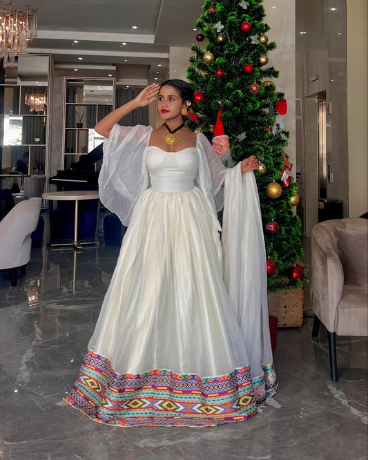 A woman wearing a white  Ethiopian Dress  with sheer draped sleeves and a colorful geometric patterned hem, standing in a modern indoor setting with a decorated Christmas tree in the background.