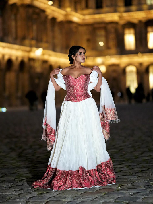 A woman stands on a black staircase wearing a traditional-style cream Ethiopian Dress with puffed sleeves, a blue sash, and gold trim, against a white wall.