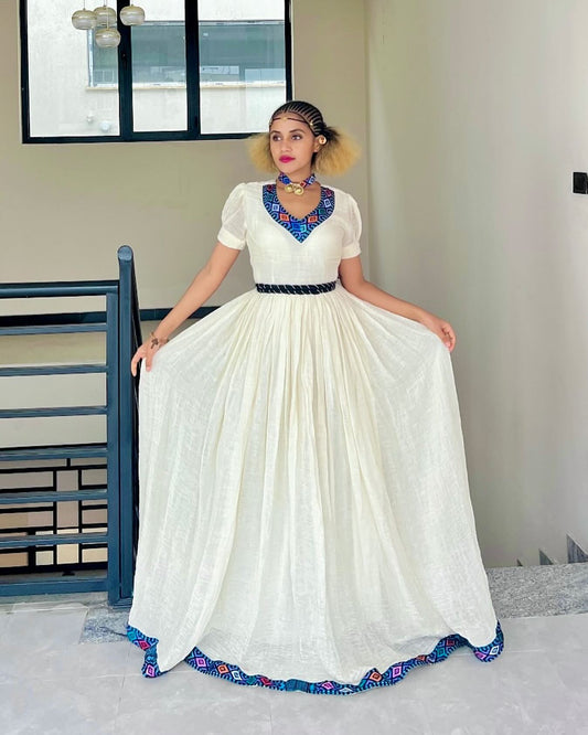 A woman modeling a long white maxi Ethiopian Dress with a black waist belt and vibrant blue, purple, and orange geometric embroidery on the neckline and hem, standing in an indoor setting.