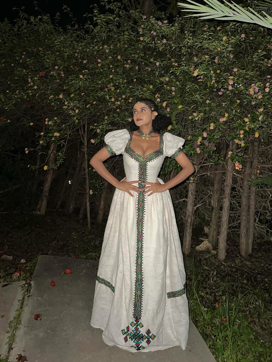 A woman wearing a long white traditional-style Habesha kemis with green and red embroidered details, standing outdoors at night in front of flowering bushes.