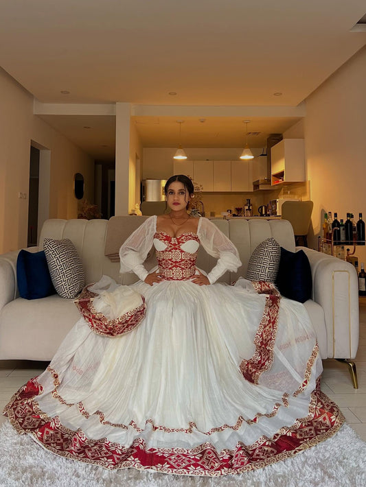 A woman seated on a cream sofa in a living room, wearing an ornate white traditional Ethiopian Dress with red embroidery and a matching red-and-white embroidered headpiece. The room has warm lighting, a sofa with blue and patterned pillows, and a kitchen area in the background.