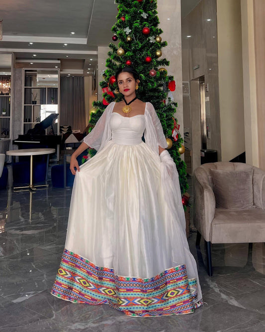 A woman wearing a white  Ethiopian Dress with sheer draped sleeves and a colorful geometric patterned hem, standing in a modern indoor setting with a decorated Christmas tree behind her.