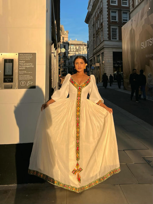 A woman stands on a black staircase wearing a traditional-style cream Ethiopian Dress with puffed sleeves, a blue sash, and gold trim, against a white wall.