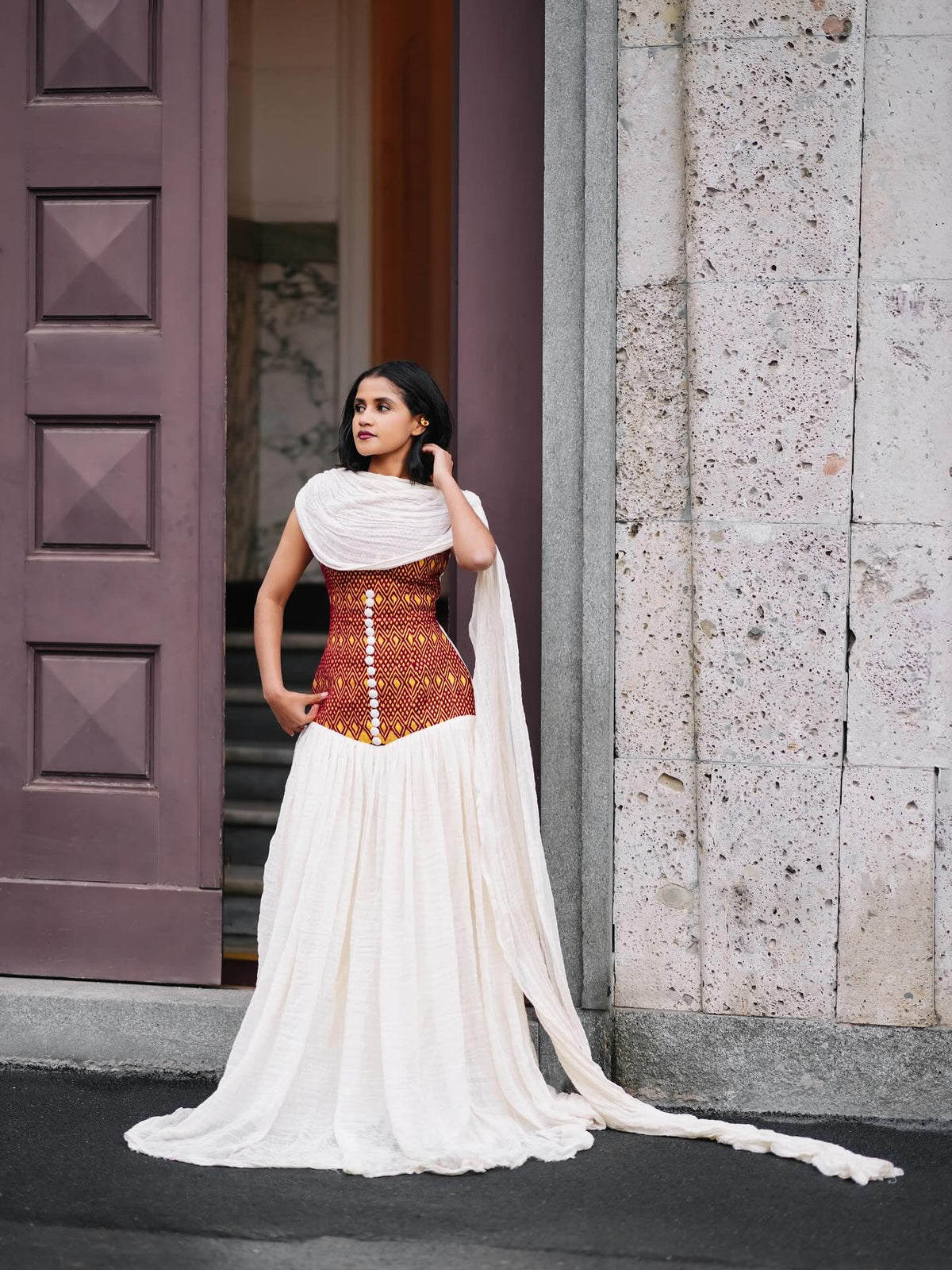 A woman wearing a white and rust-orange embroidered habesha kemis with a long flowing train, standing outdoors in front of a stone wall and a dark wooden door.