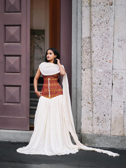A woman wearing a white and rust-orange embroidered habesha kemis with a long flowing train, standing outdoors in front of a stone wall and a dark wooden door.