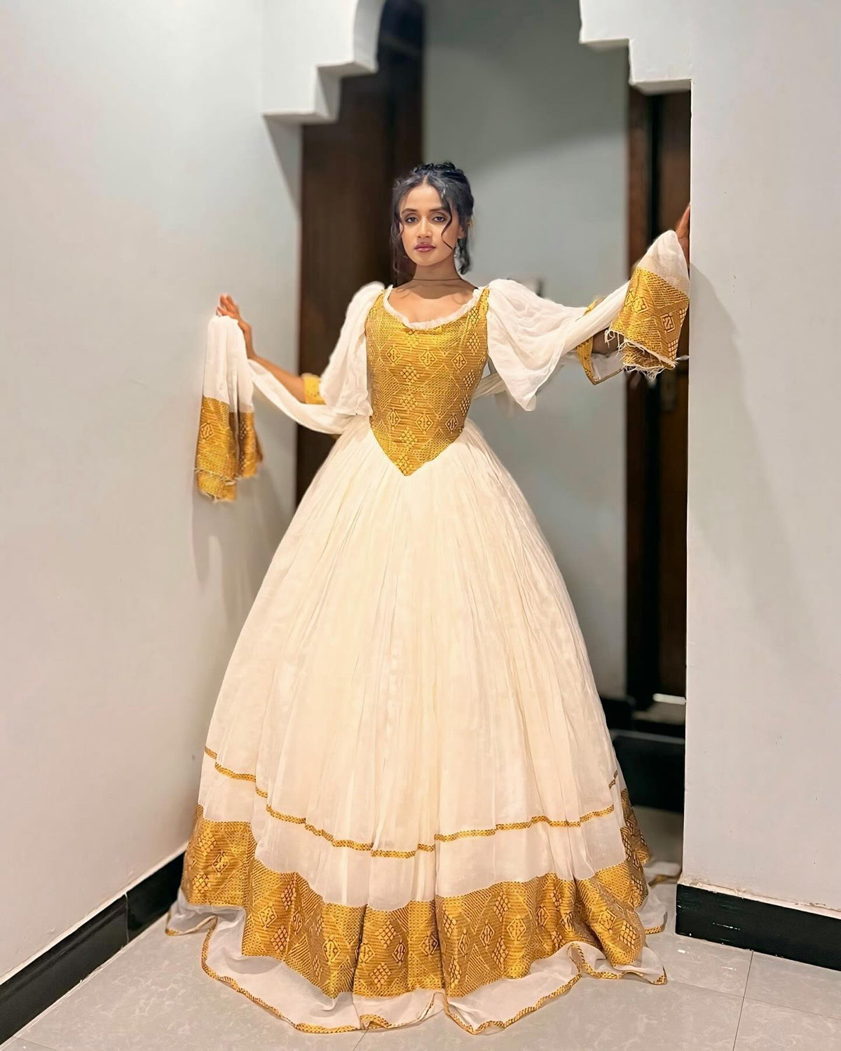 A woman modeling a traditional Ethiopian Dress with a cream-colored skirt, a gold embroidered bodice, and gold-trimmed sheer sleeves, standing in a doorway.