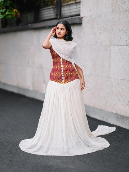 A woman stands outdoors against a light stone wall wearing a long white habesha kemis with a fitted rust-red and gold patterned bodice and a flowing white skirt with a train. She also wears a white draped shawl.