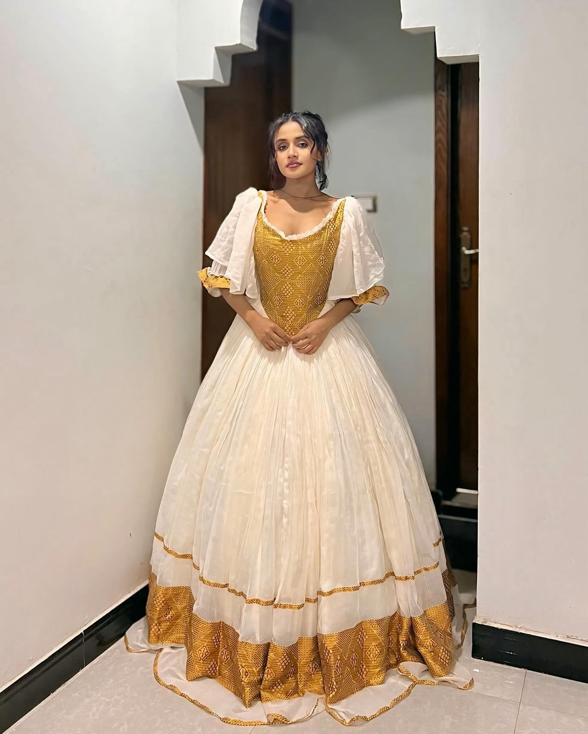 A woman wearing a traditional Ethiopian Dress with a cream-colored skirt, gold embroidered bodice, and white sheer flutter sleeves, standing in an indoor hallway.