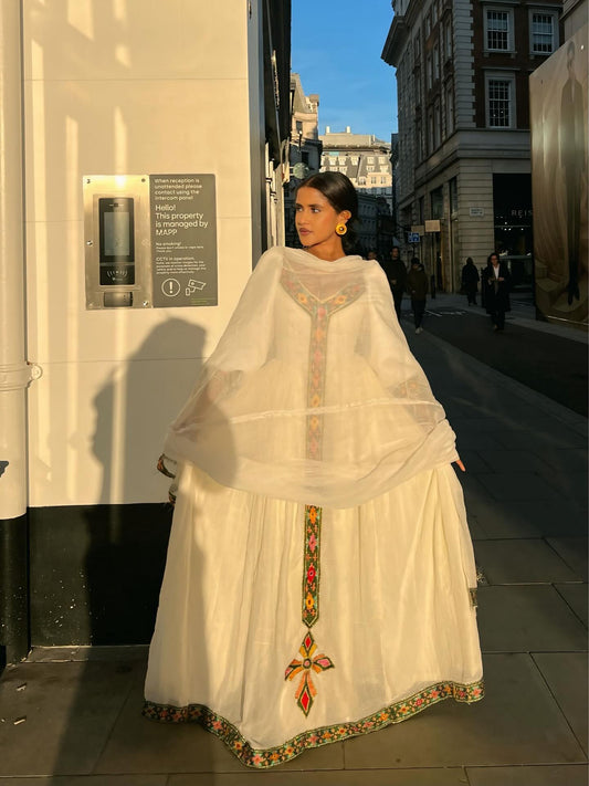 A woman stands on a black staircase wearing a traditional-style cream Ethiopian Dress with puffed sleeves, a blue sash, and gold trim, against a white wall.