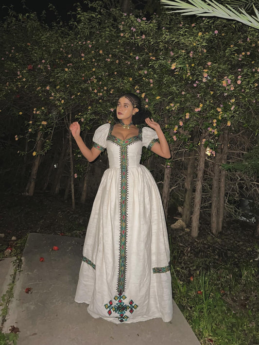 A woman wearing a long white Habesha kemis with colorful embroidered details stands outdoors at night in front of leafy bushes.