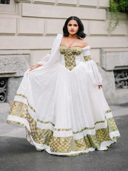 A woman modeling a white off-the-shoulder traditional Ethiopian Dress with ornate gold and green patterned trim, standing outdoors against a stone building facade.