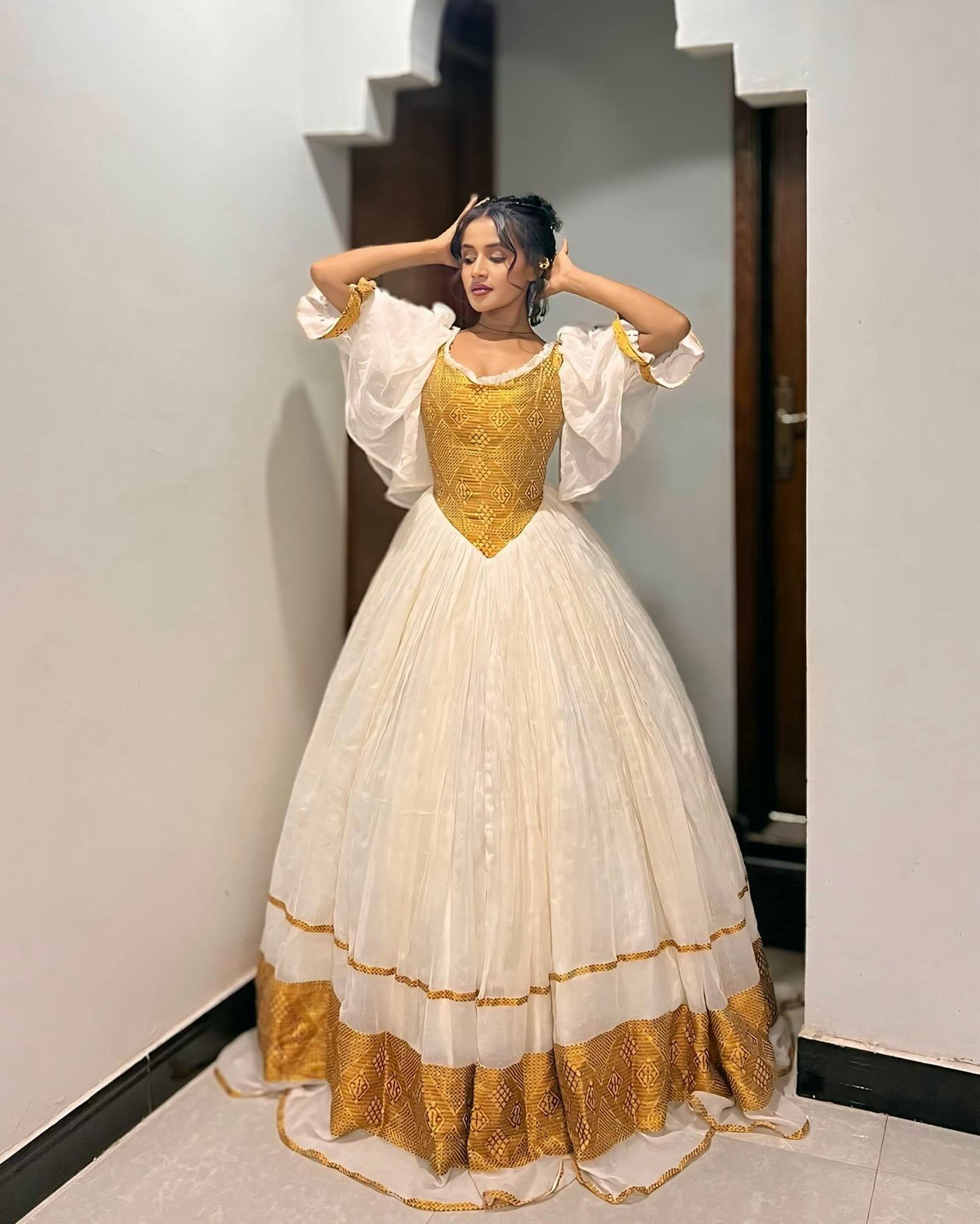 A woman wearing a Ethiopian Dress with a gold embroidered bodice and gold patterned hem, posing in an indoor hallway.