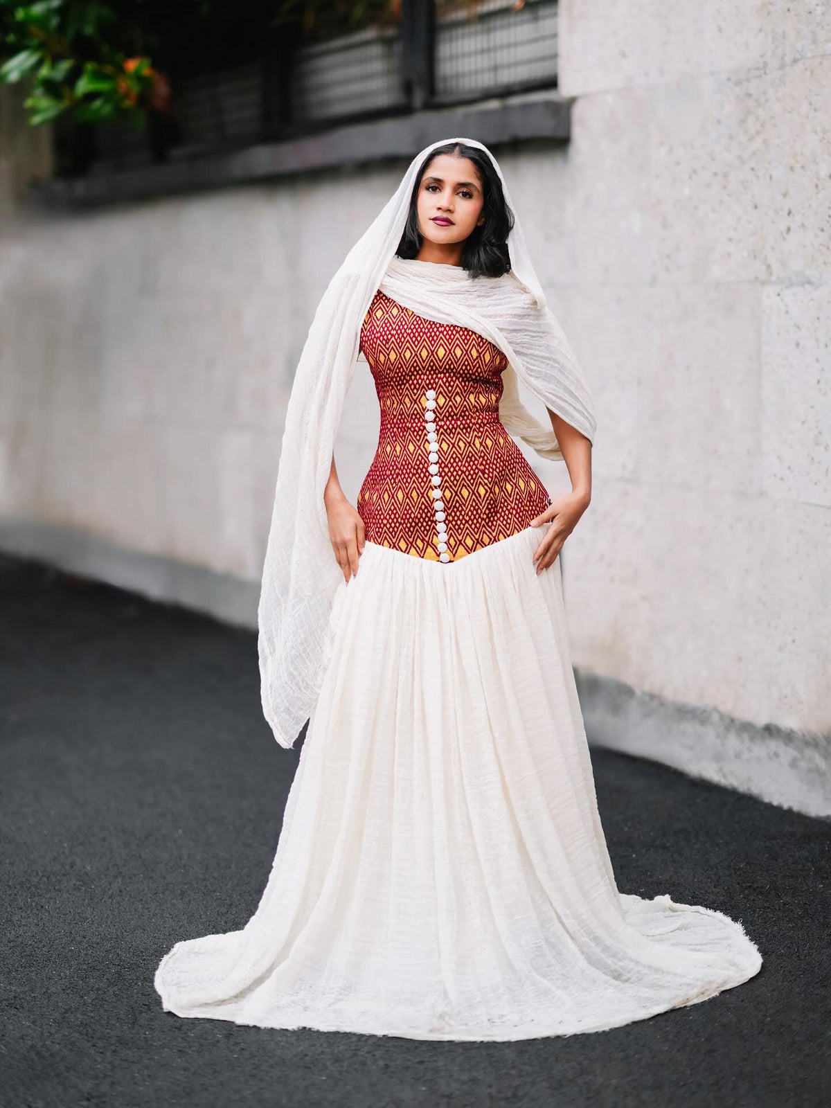 A woman stands outdoors wearing a habesha kemis with a fitted rust-red embroidered bodice, a long white skirt, and a sheer white draped head covering. The background shows a light stone wall and dark pavement.