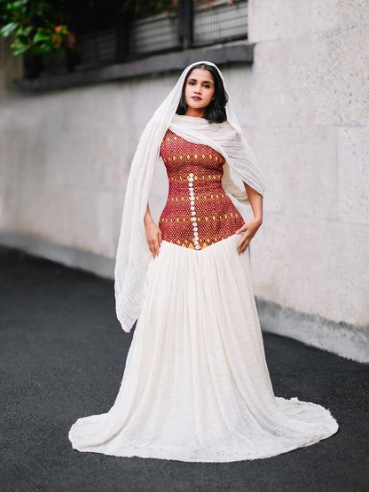 A woman stands outdoors wearing a habesha kemis with a fitted rust-red embroidered bodice, a long white skirt, and a sheer white draped head covering. The background shows a light stone wall and dark pavement.