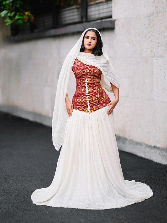 A woman stands outdoors wearing a habesha kemis with a fitted rust-red embroidered bodice, a long white skirt, and a sheer white draped head covering. The background shows a light stone wall and dark pavement.