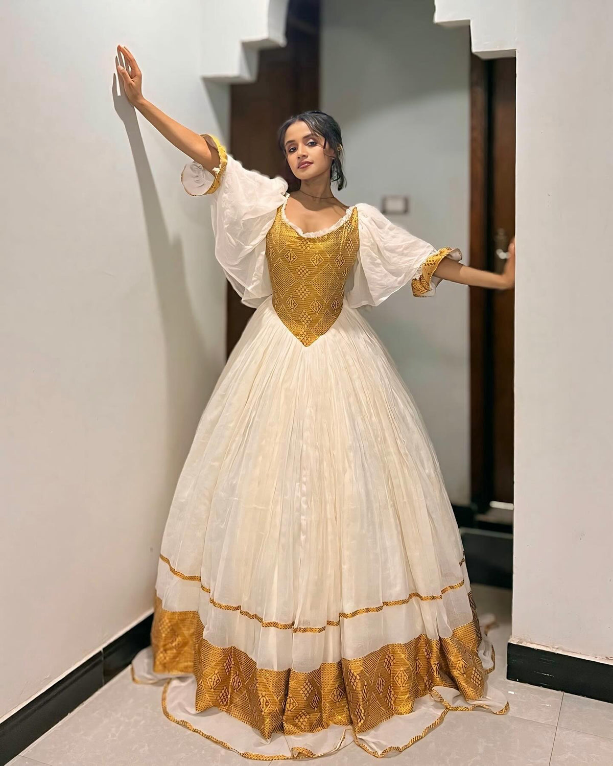 A woman modeling a traditional Ethiopian Dress with a cream-colored tulle skirt, a gold embroidered bodice, and gold-trimmed sheer sleeves, posing in an indoor hallway.