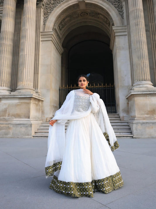 A woman wearing a traditional white habesha Dress with green embroidered details stands on stone steps in front of a grand stone building entrance with a black metal gate.