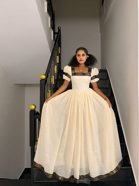 A woman stands on a black staircase holding out a voluminous off-white Ethiopian Dress with puffed sleeves and ornate gold detailing along the neckline and hem.