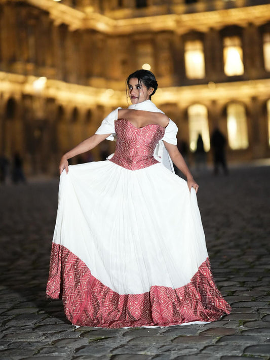 A woman stands on a black staircase wearing a traditional-style cream Ethiopian Dress with puffed sleeves, a blue sash, and gold trim, against a white wall.
