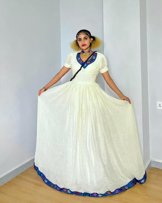 A woman modeling a traditional white or cream-colored long Ethiopian Dress with a full skirt, short sleeves, and a blue embroidered neckline and hem, standing in a minimalist room with light gray walls and a wooden floor.