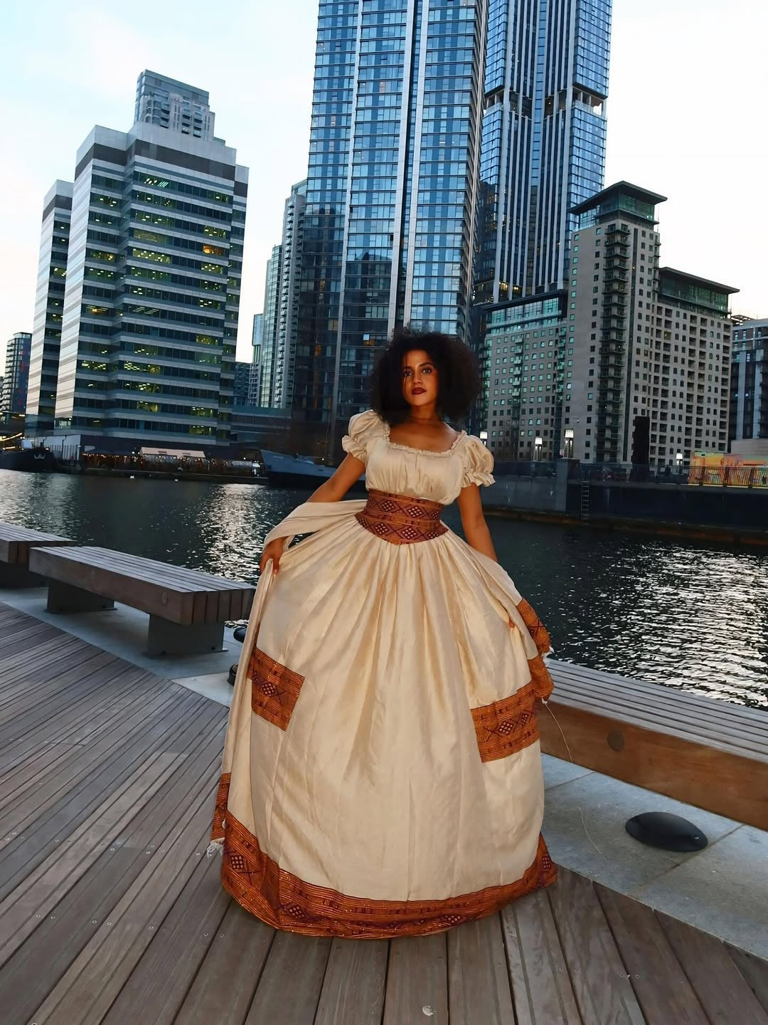 Woman in a Brown Habesha Libs standing on a dock with city skyline in the background