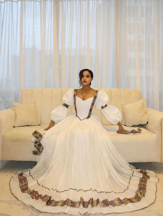 Woman in a white Gold Floral Habesha Dress with decorative trim sitting on a beige sofa.