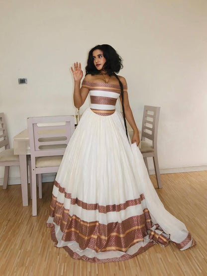 Woman in a white and brown traditional Ethiopian Traditional Dress standing in a room with wooden floor and chairs.