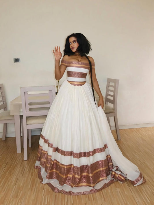 Woman in a white and brown traditional Ethiopian Traditional Dress standing in a room with wooden floor and chairs.