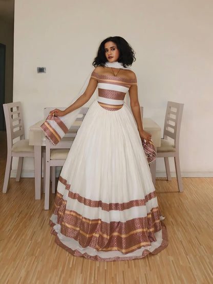 Woman in a white and brown Ethiopian Traditional Dress standing in a room with wooden flooring and chairs.