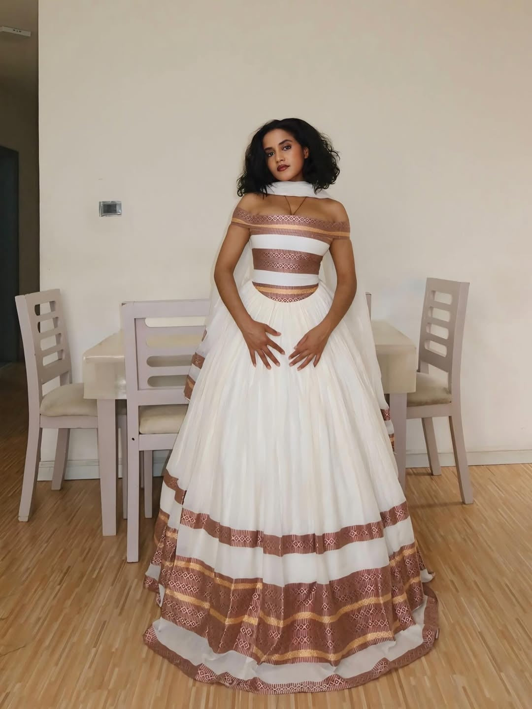 Woman in a white and brown Ethiopian Traditional Dress standing in a room with chairs and a table.