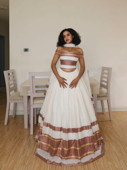 Woman in a white and brown Ethiopian Traditional Dress standing in a room with chairs and a table.