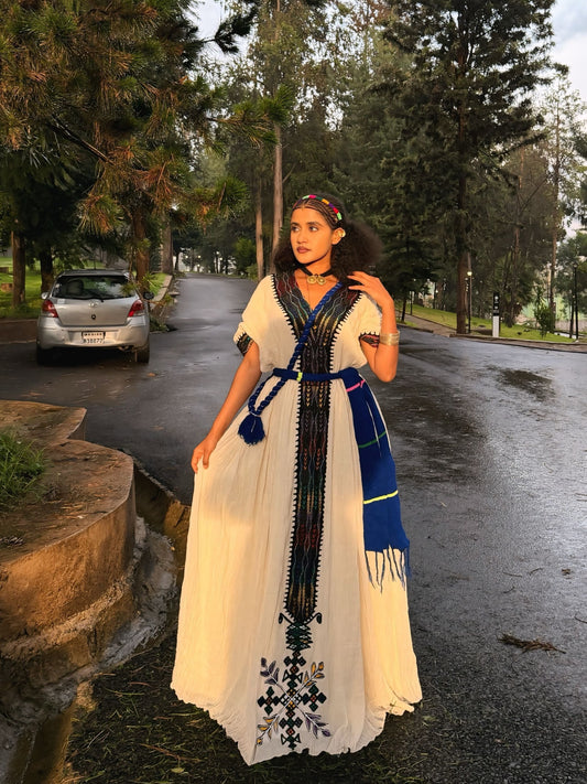 A woman stands on a wet road outdoors wearing a long cream Ethiopian Dress with blue and black embroidered details, a blue sash, and a blue fringed scarf. Trees and a parked silver car are visible in the background.