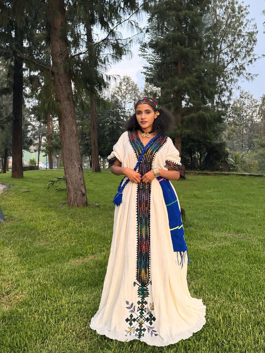 A woman wearing a long cream-colored Ethiopian Dress with colorful embroidered details and a blue sash stands outdoors on a grassy lawn with trees in the background.