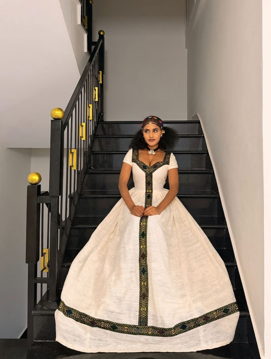 A young girl stands on a black staircase wearing a traditional Habesha Kemis with a voluminous white skirt, short sleeves, and intricate black and gold embroidery.
