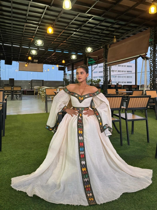 A woman models a traditional off-the-shoulder white Traditional Ethiopian Dress with colorful geometric embroidered details along the neckline, waist, and center front panel, set against an outdoor event space with tables and artificial grass.