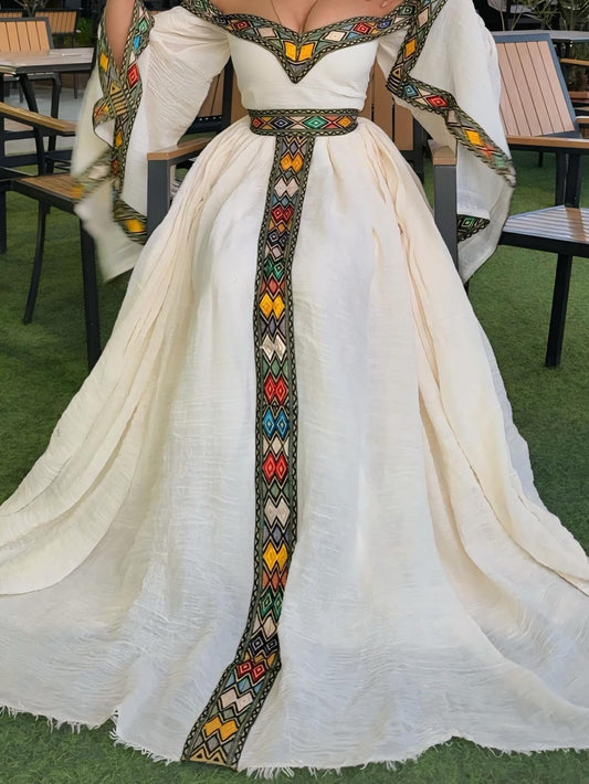 A woman wearing a voluminous off-white or cream historical-style Traditional Ethiopian Dress with colorful geometric embroidered trim along the neckline, sleeves, waist, and center front panel, photographed outdoors on artificial grass with patio chairs visible in the background.
