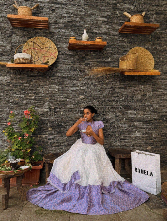 Woman in a white and purple  Habesha dress sitting on the ground with wicker baskets and a 'RAHELA' bag in the background.