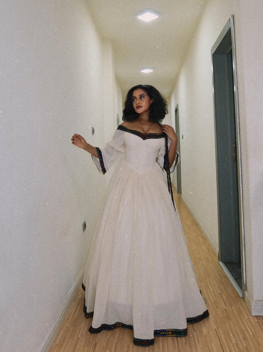 A woman wearing a white off-the-shoulder ball  Habesha dress with black trim stands in a hallway with light wood floors and off-white walls.