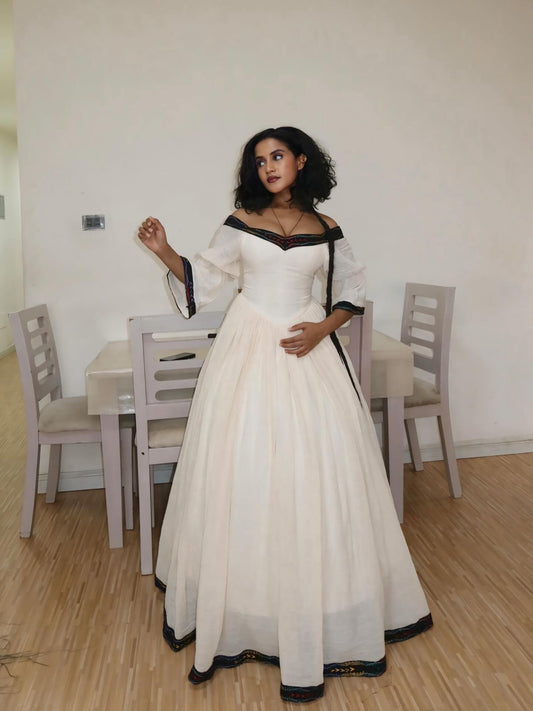 A woman wearing a long off-white ball  Habesha dress with black trim and off-the-shoulder sleeves, standing in a room with a wooden floor and dining chairs in the background.