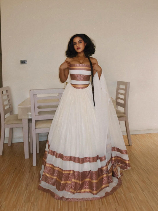 A woman wearing a traditional white Habesha Kemis with metallic bronze and gold patterned borders, standing in a room with wooden floors and chairs.