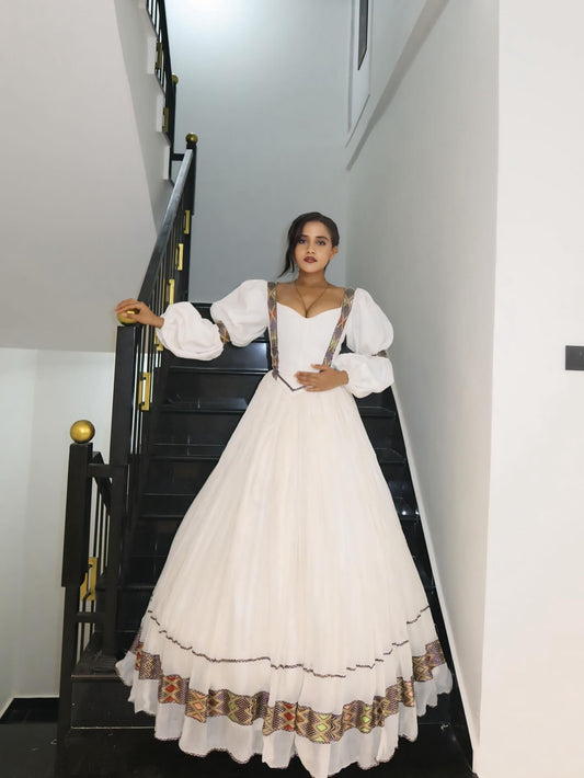 A woman wearing a white traditional Habesha Kemis with patterned suspenders and a full skirt, posing on a black staircase indoors.